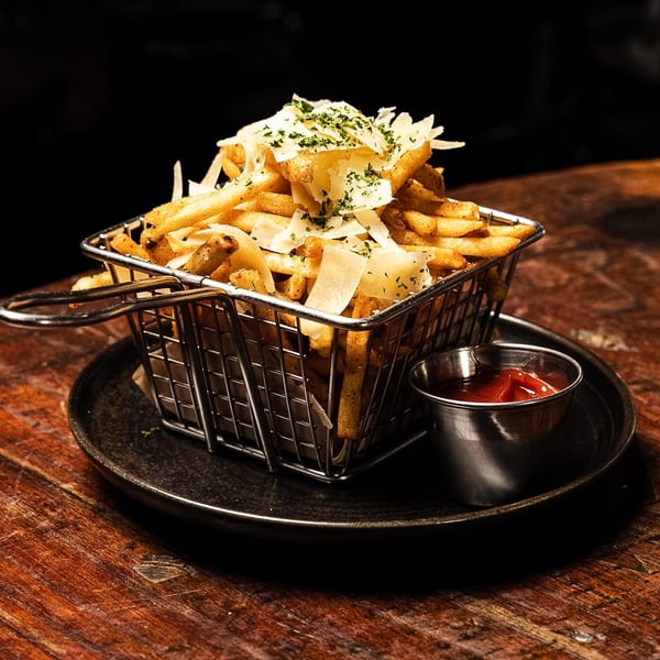 Crispy french fries topped with shaved parmesan and parsley, served with ketchup in a metal basket.