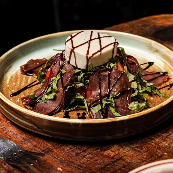 A gourmet dish with sliced meat, greens, and a block of cheese drizzled with balsamic glaze.