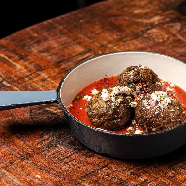 Three meatballs in red sauce, topped with feta and herbs, in a small skillet on a wooden table.