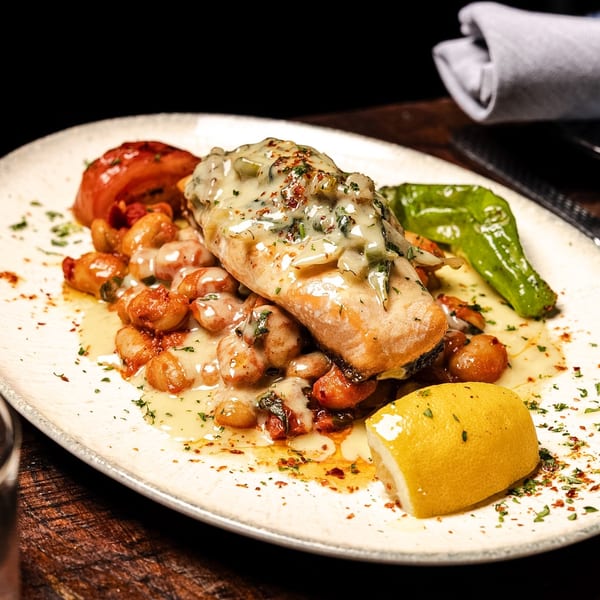 Salmon fillet with creamy sauce, beans, roasted tomato, green pepper, and lemon on a plate.